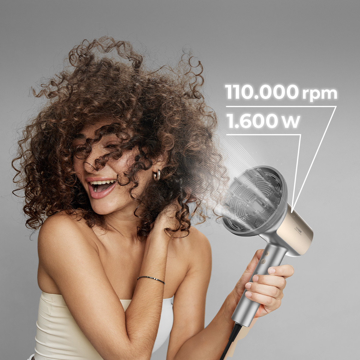 Mujer con rizos voluminosos seca su pelo con secador plateado, gráfico de texto indica 110.000 rpm y 1.600 w.