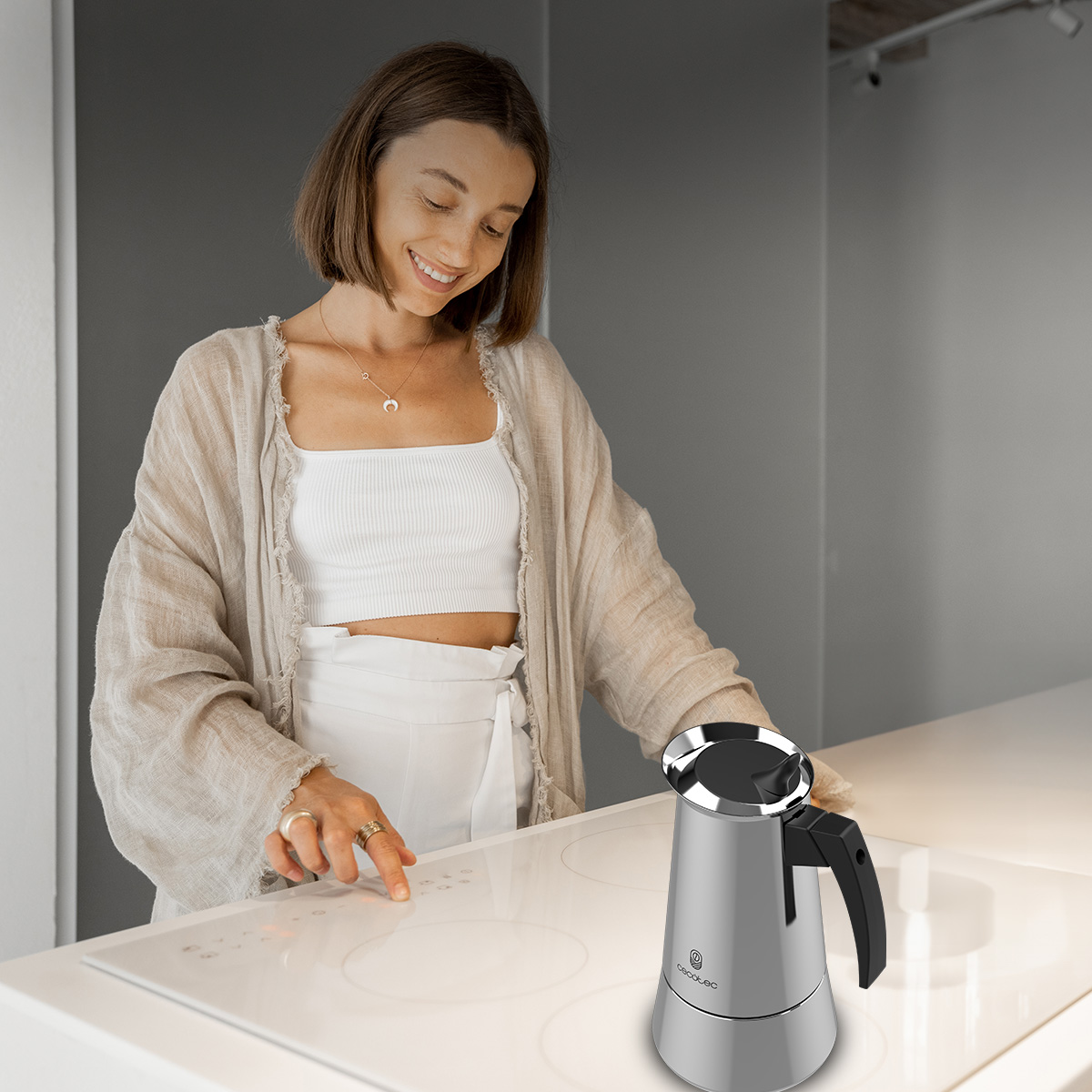 Una mujer sonriente en blanco y beige toca los controles de una placa de inducción blanca junto a una cafetera.