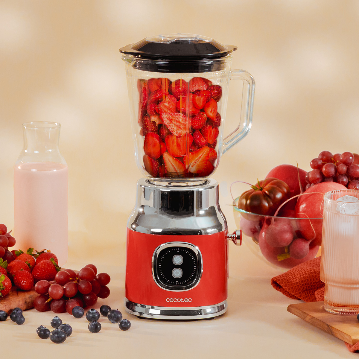 Licuadora roja llena de fresas, rodeada de leche, fruta y un vaso de batido sobre fondo beis.