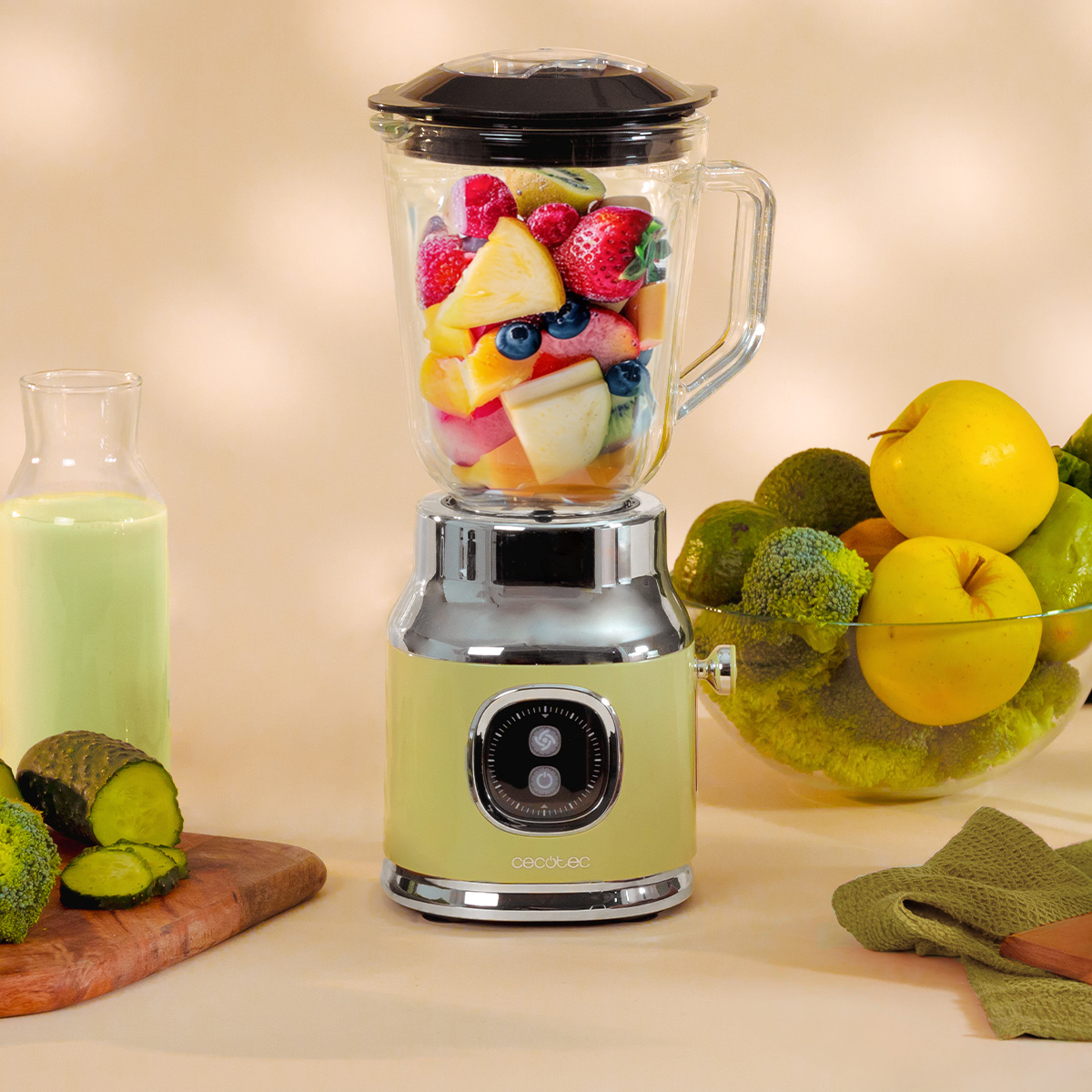 Una licuadora verde pastel llena de fruta mezclada está al lado de una botella de batido, manzanas y brócoli.