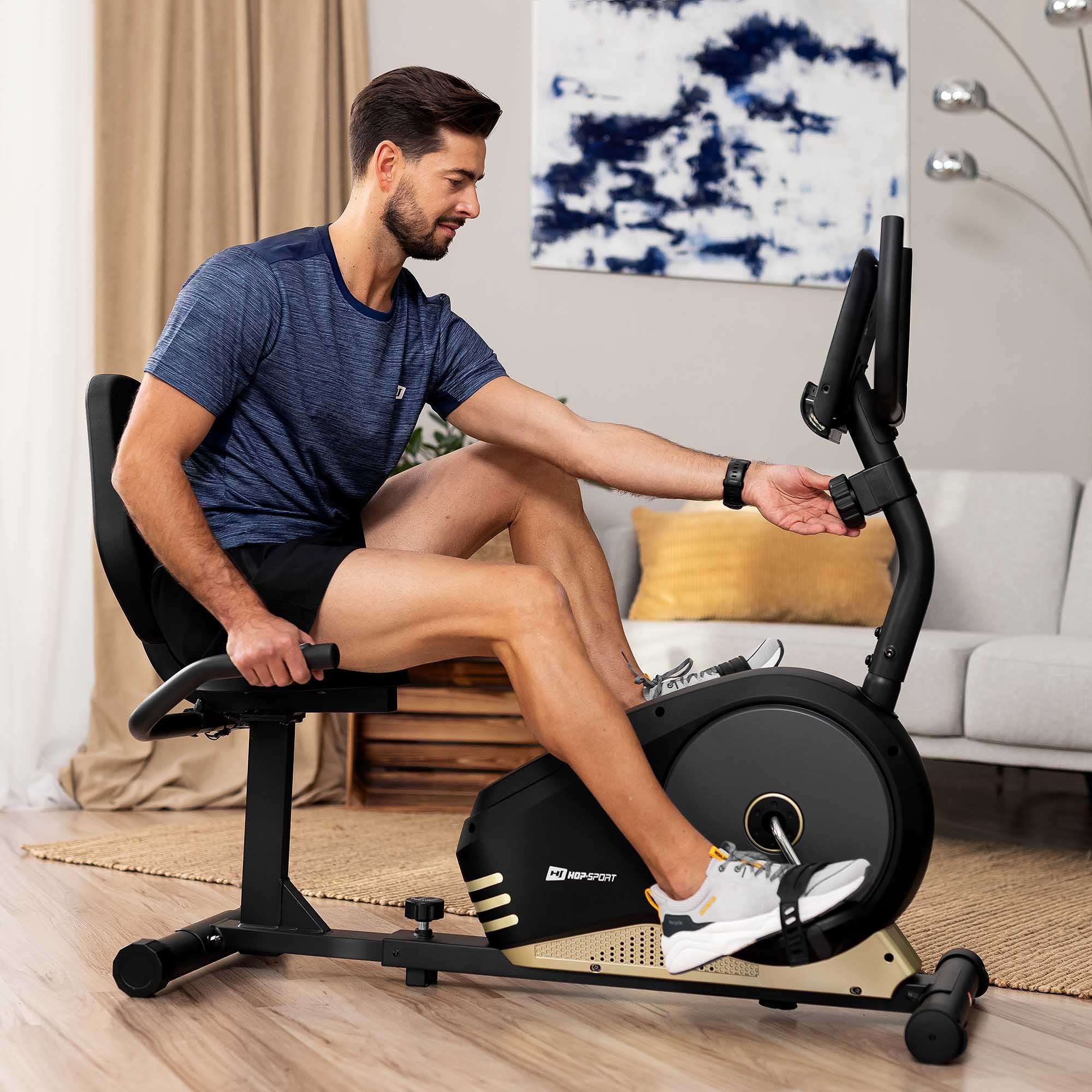 Ein Mann sitzt in einem modernen Wohnzimmer auf einem schwarzen Heimtrainer und stellt das Display ein.