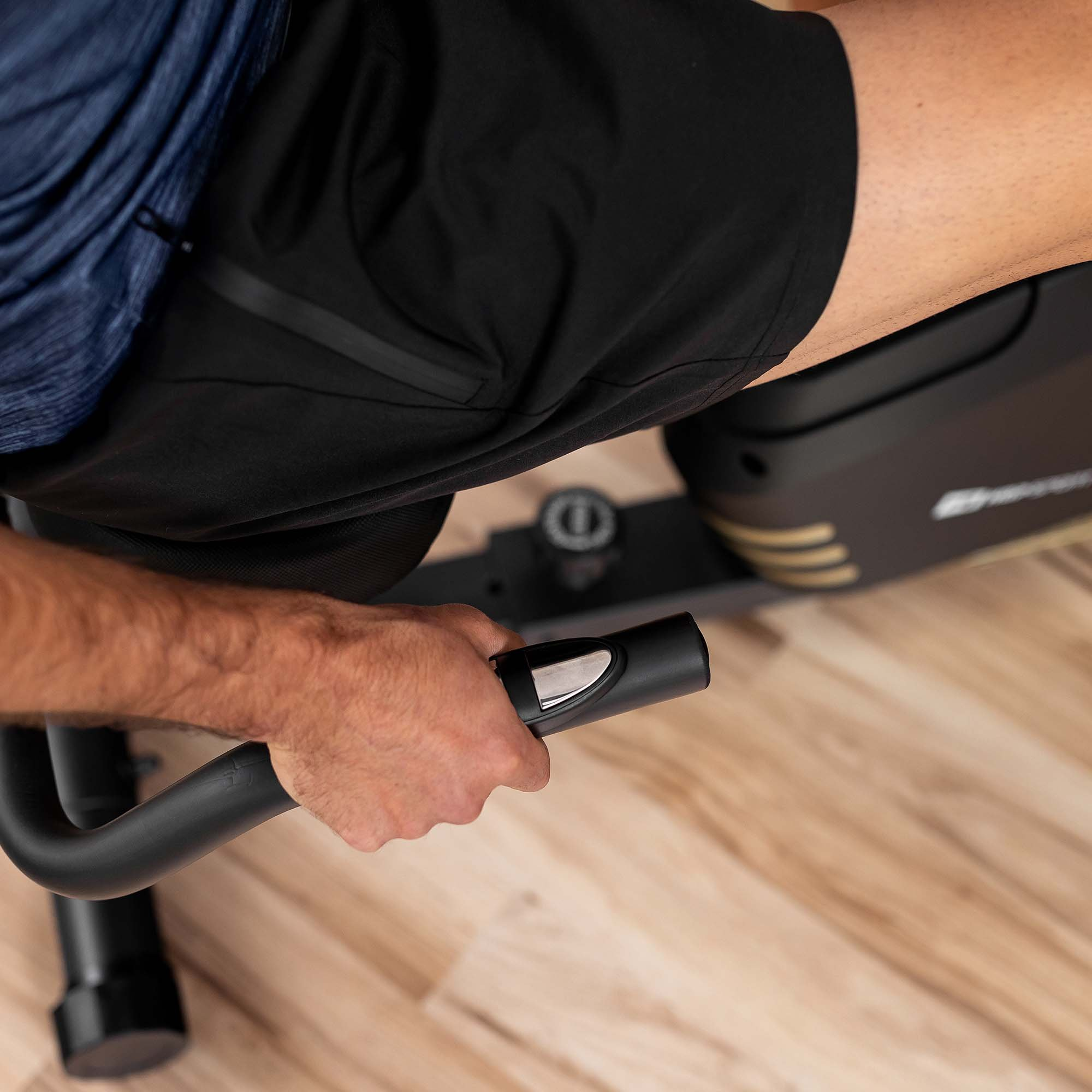 Eine Person in schwarzen Shorts und blauem Hemd trainiert auf einem Hometrainer und hält den Lenker auf einem Holzboden.