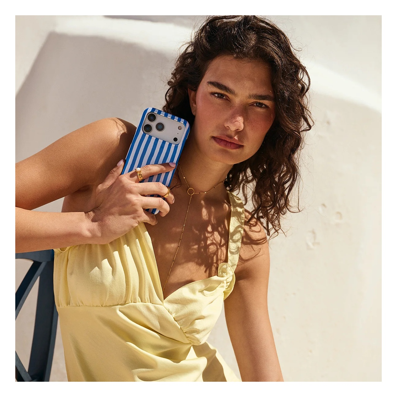 A woman with curly hair wears a yellow dress, holding a blue and white striped phone case against a white wall.