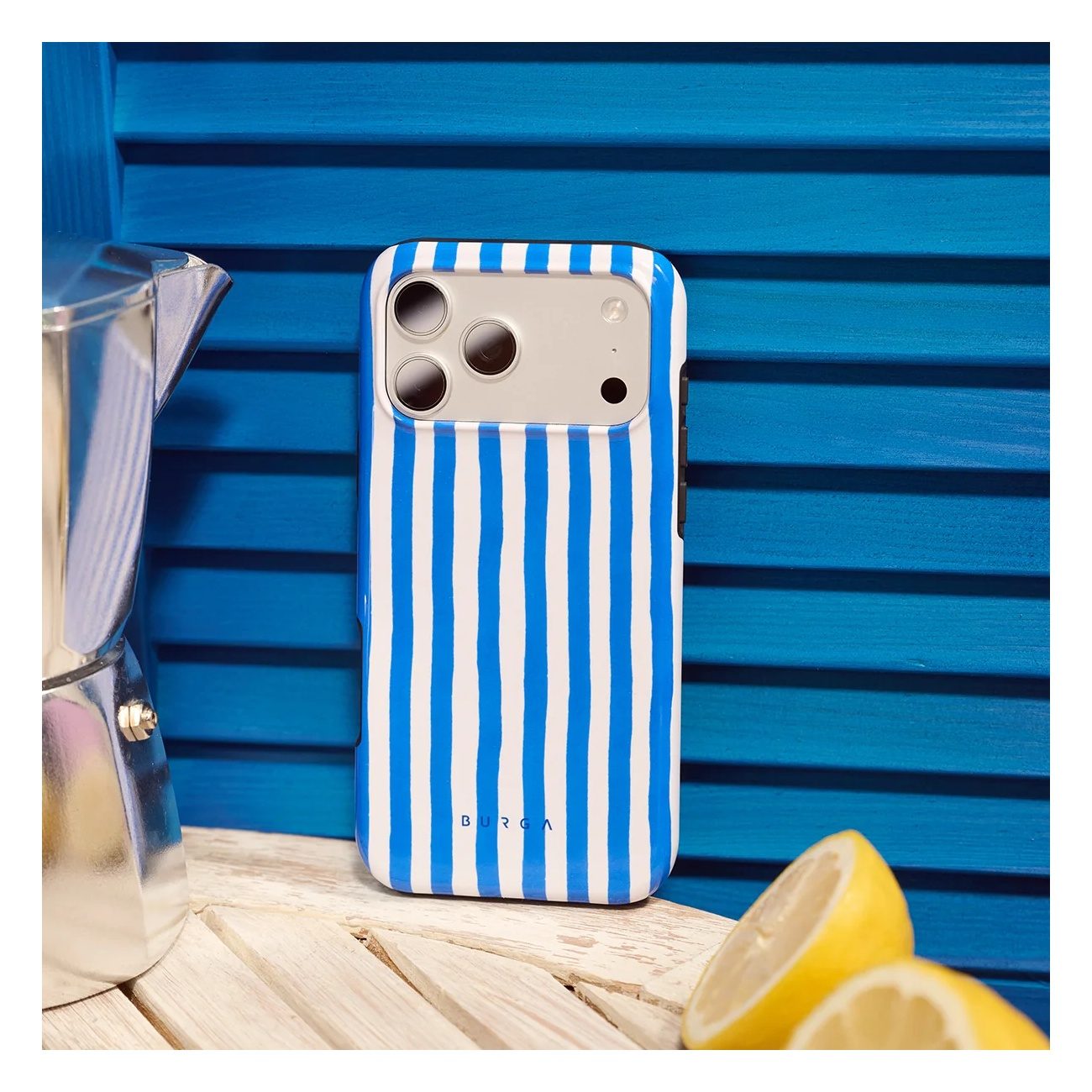A blue and white striped phone case sits on a wooden surface beside a metal coffee maker and cut lemons.
