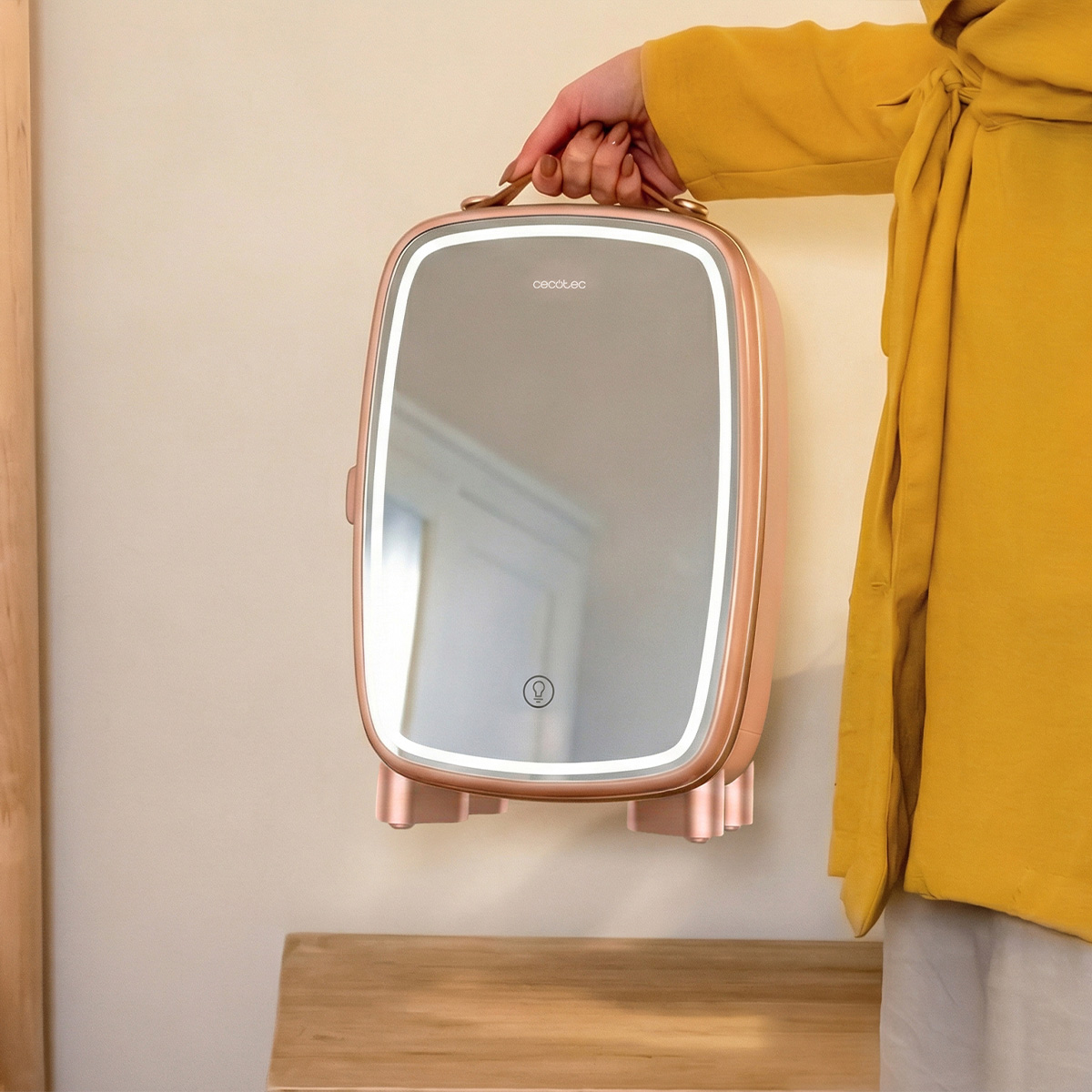 Una mano con cappotto giallo regge una valigetta per trucco in oro rosa con specchio illuminato.