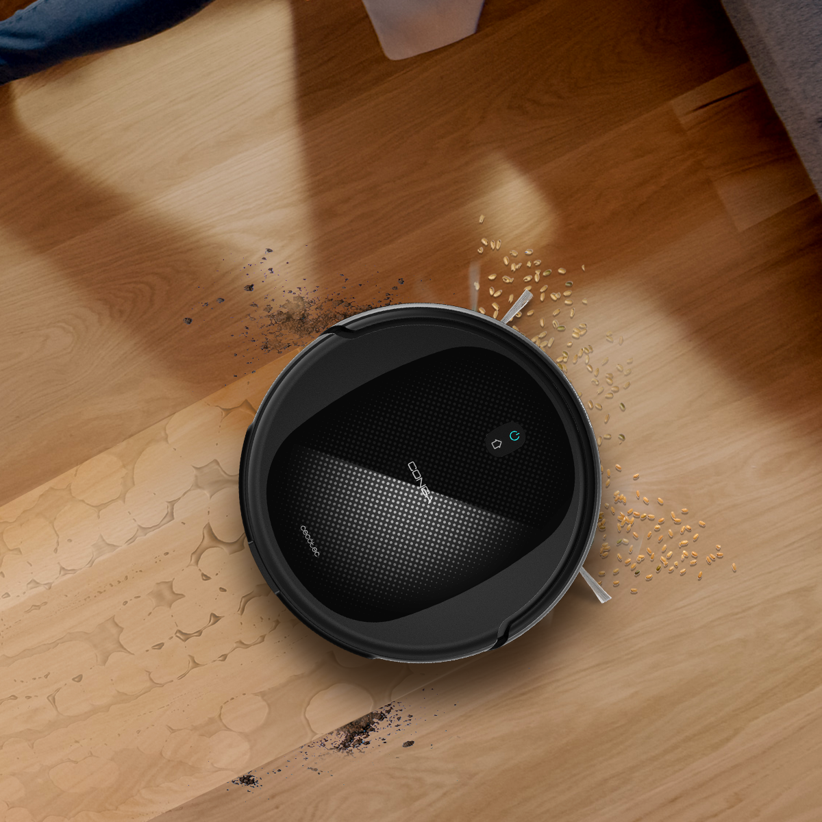 Un robot aspirapolvere nero pulisce un pavimento in legno con sporco e grano.