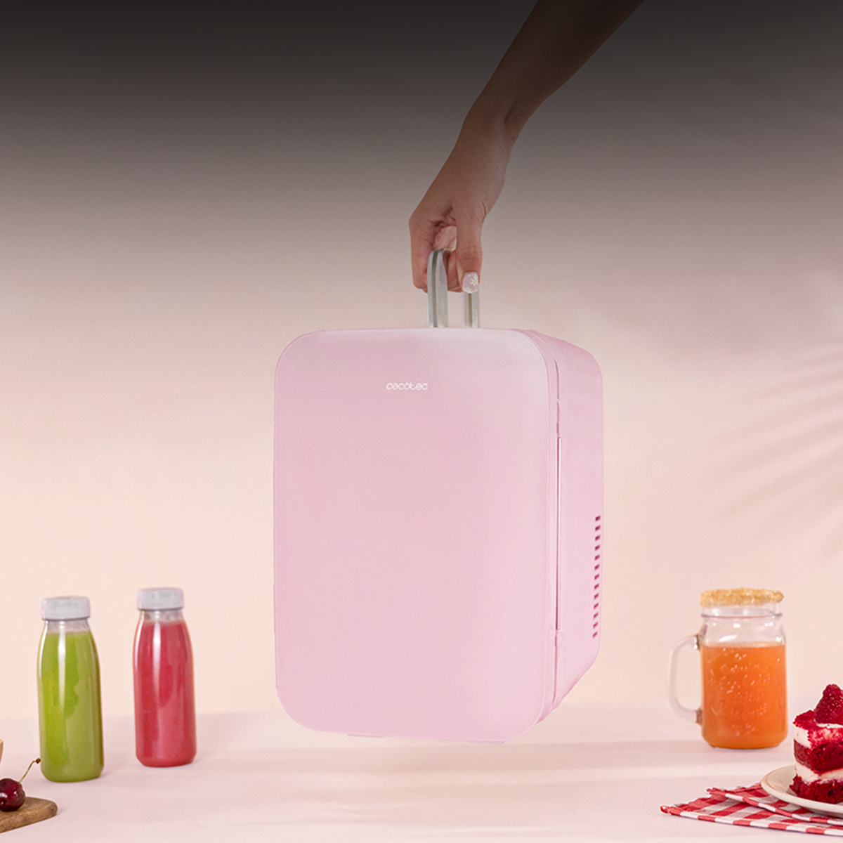 Una mano sostiene un mini refrigerador rosa. Rodeado de batidos, zumo de naranja y pastel.