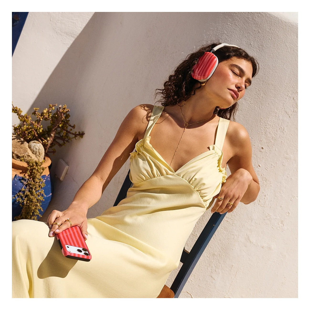 A woman in a yellow dress sits outdoors, wearing pink headphones and holding a striped phone.