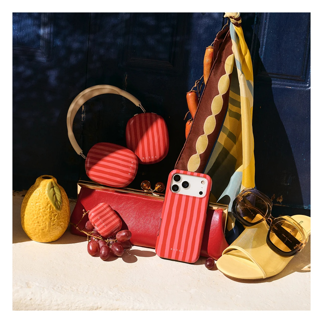 Red-and-white striped headphones, phone case, and airpod case sit with grapes and a lemon before a blue door.