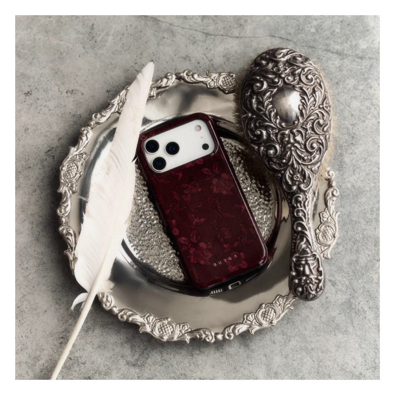 A maroon floral phone case on a silver tray with a feather and brush on a grey background.