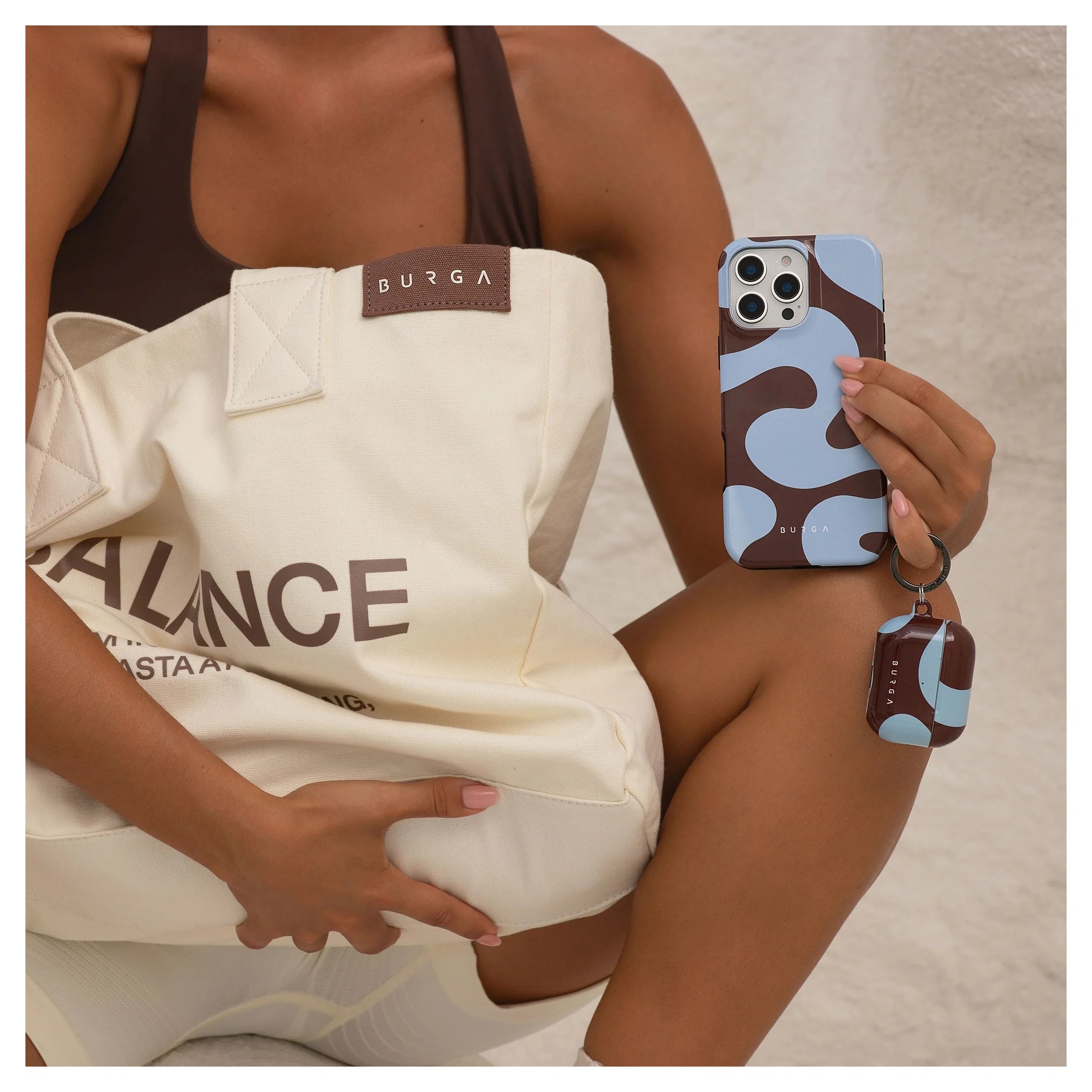A woman in brown apparel holds a beige tote bag and a blue and brown patterned smartphone.