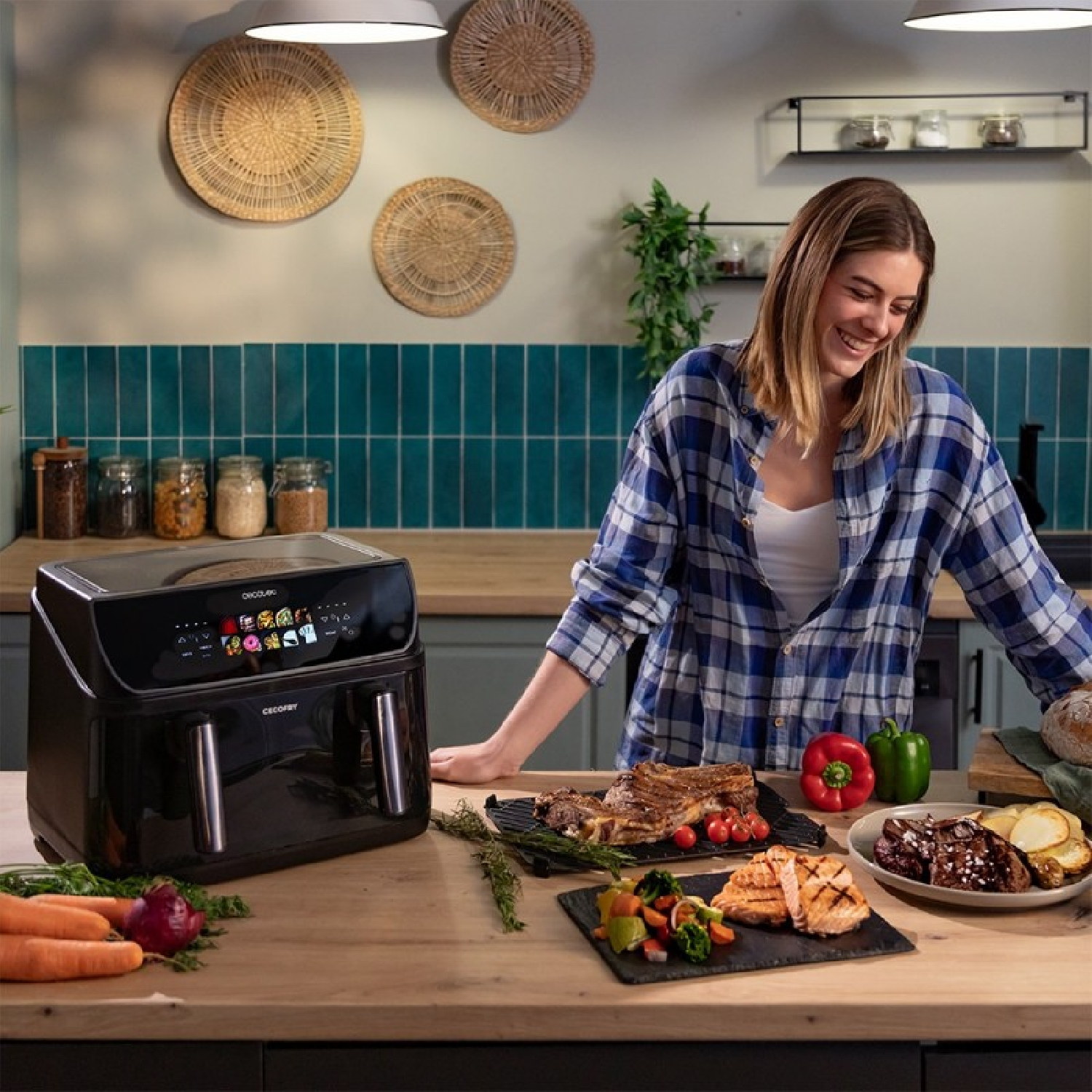 Una mujer en una cocina junto a una freidora de aire con carne, verduras y pan sobre la mesa.