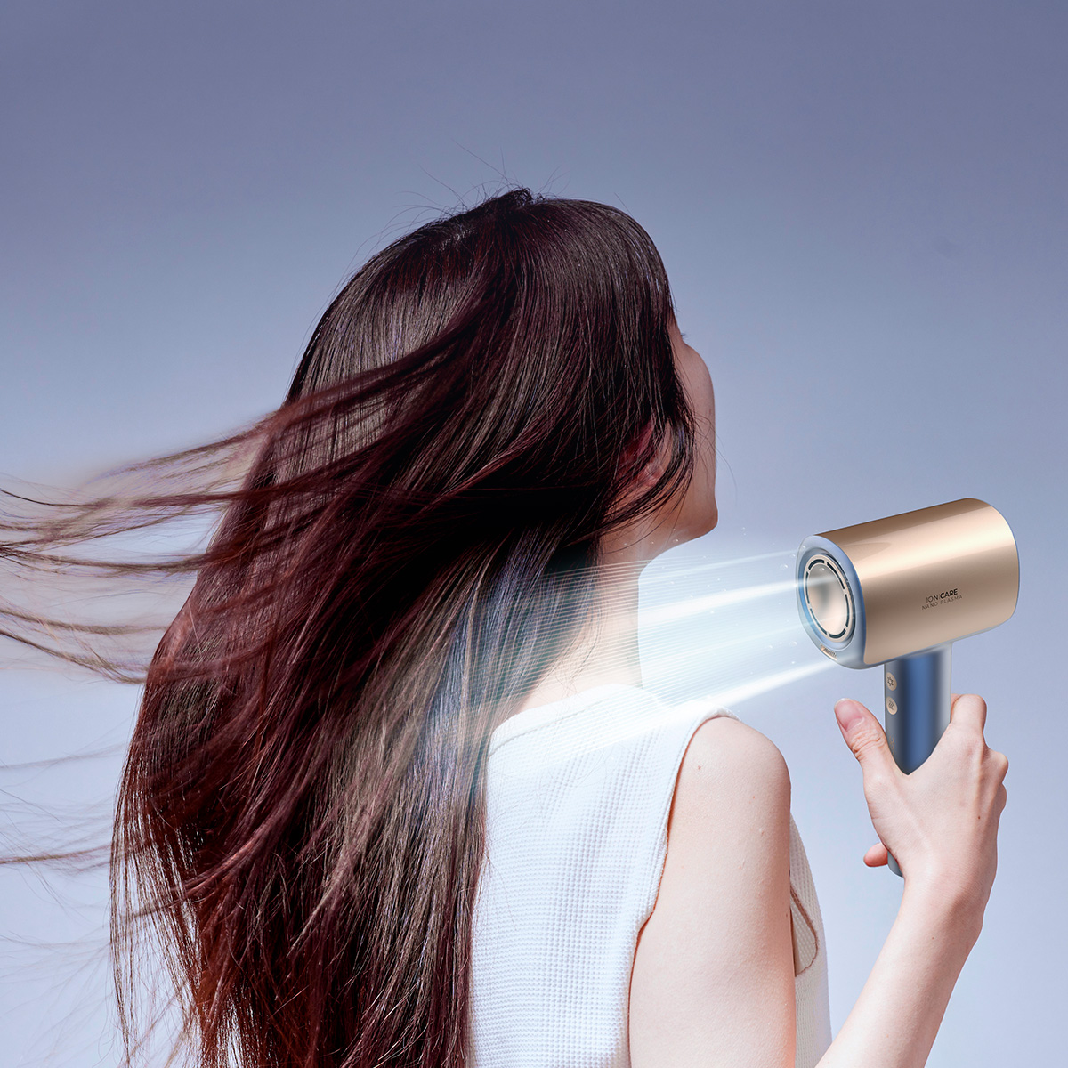 Una mujer con top blanco seca su cabello castaño largo con un secador dorado, creando movimiento.