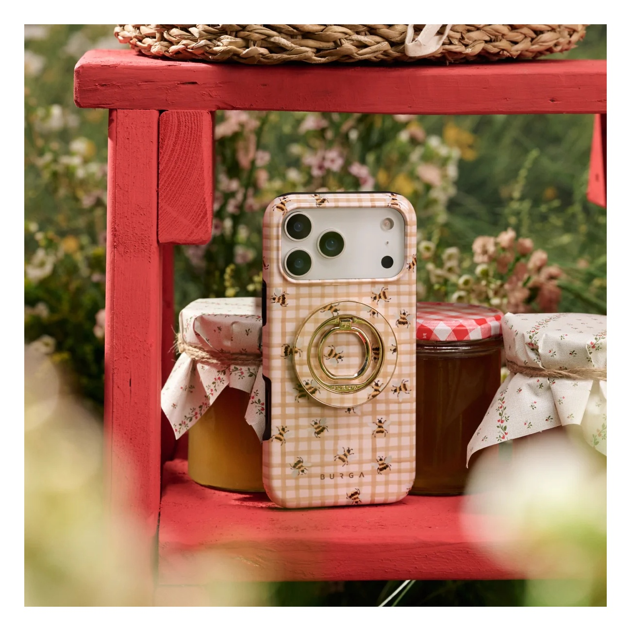 A checkered phone case with bees and a ring holder stands on a red wooden bench among jars.