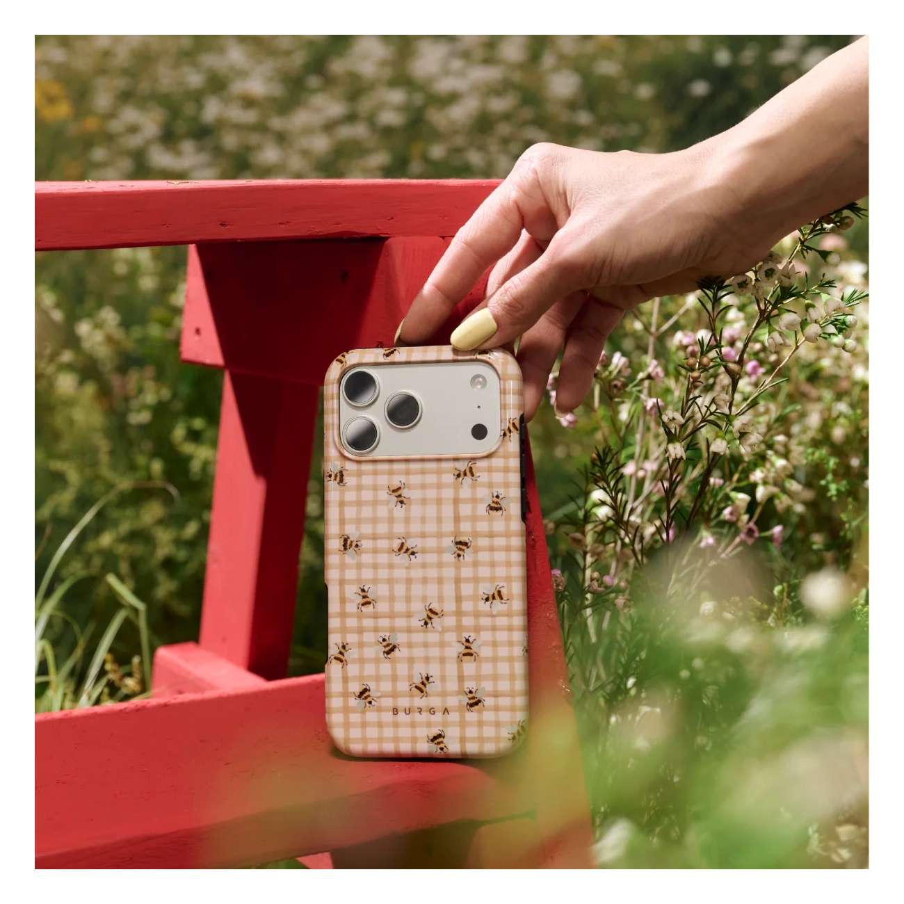 A hand holds a beige phone case with a bee pattern in front of a red wooden frame in a garden.