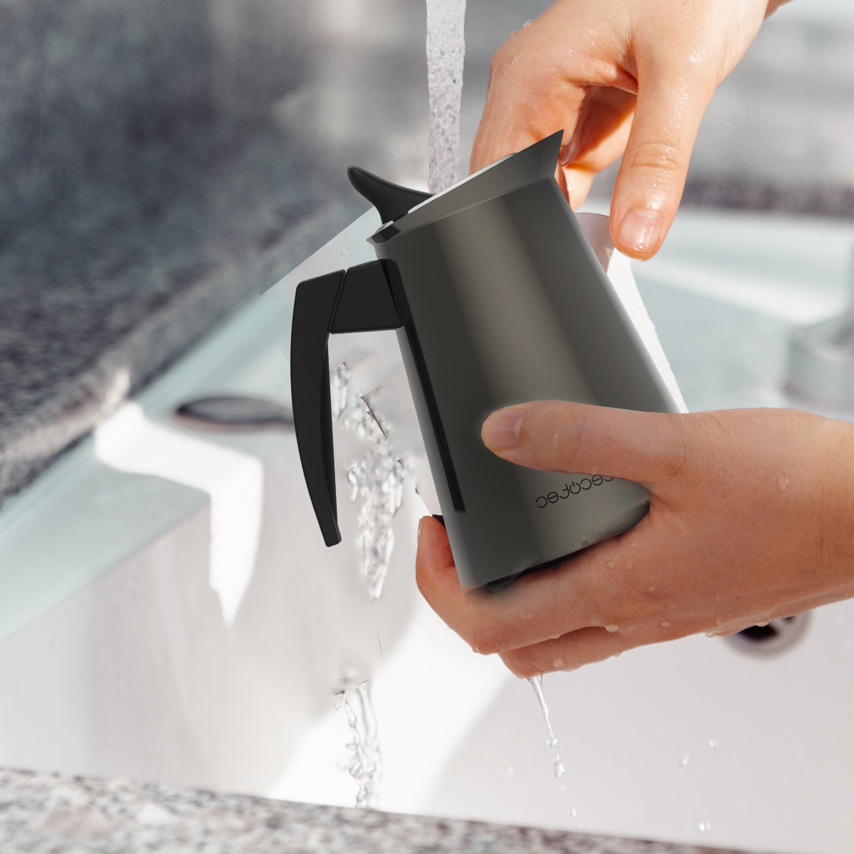 Manos lavando una cafetera gris oscura bajo agua corriente sobre un lavabo blanco.