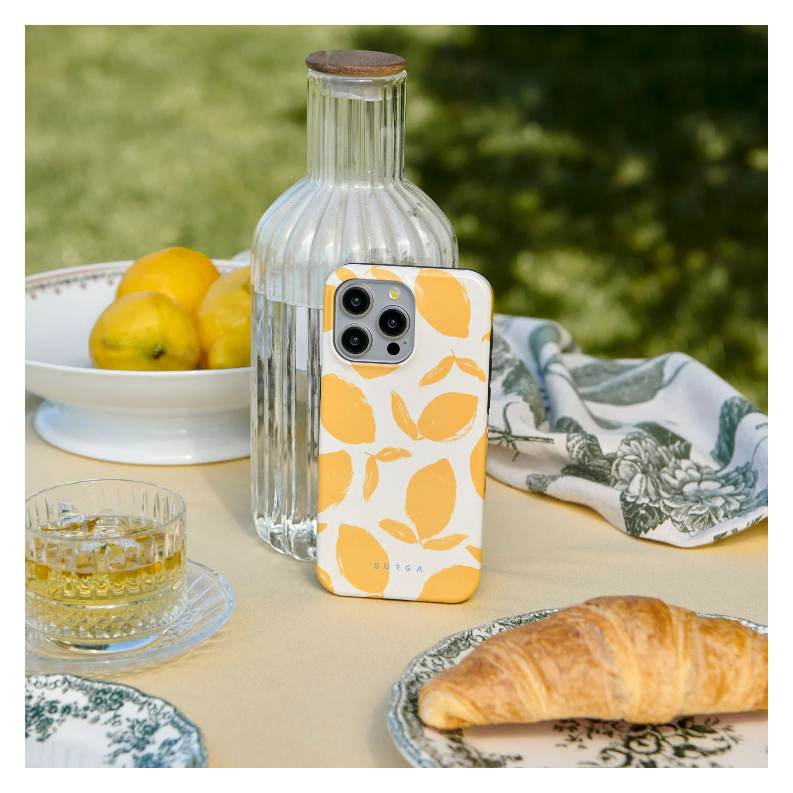 A lemon-patterned phone case stands upright on an outdoor table beside a glass bottle, a cup of tea, and a croissant.