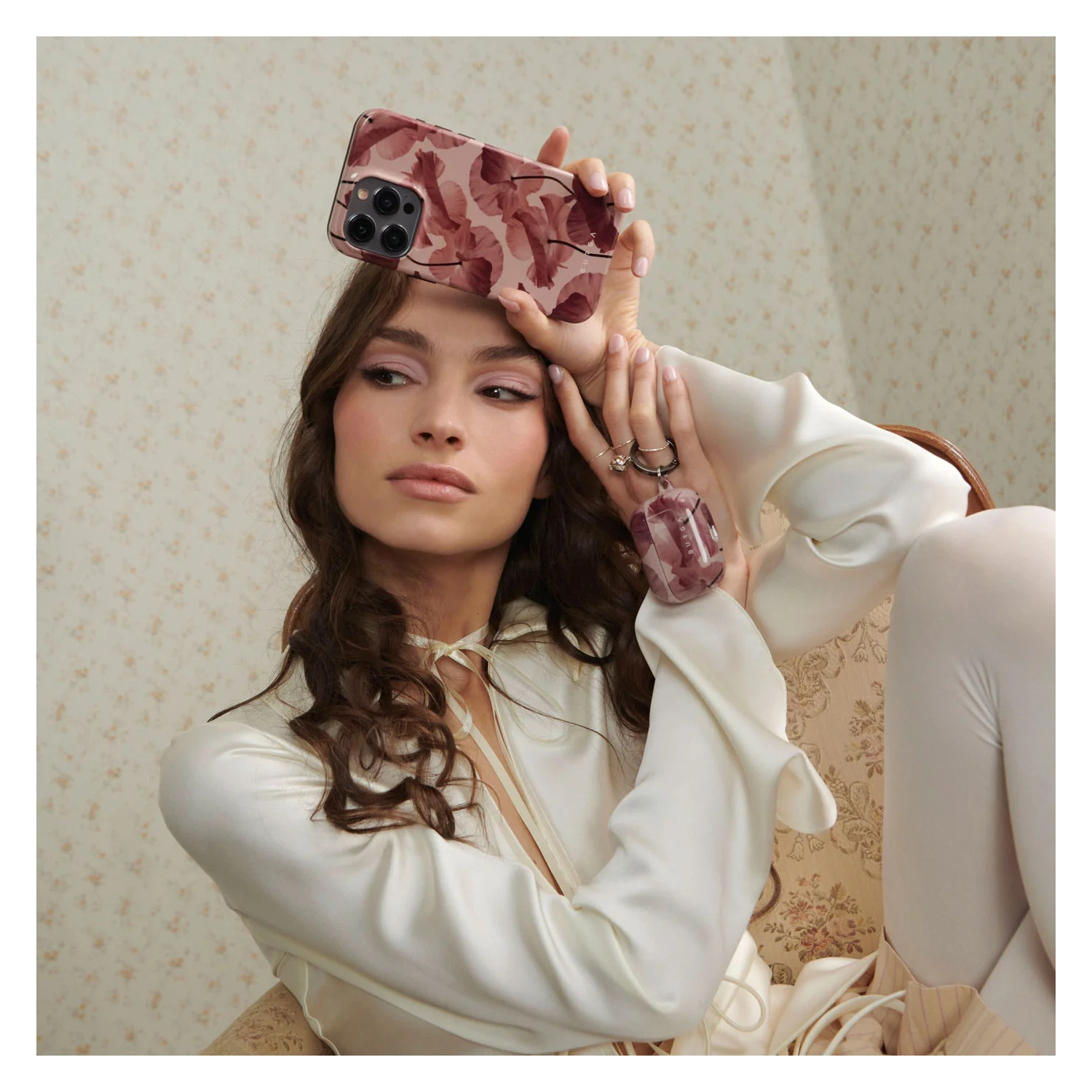 A woman in a white blouse holds a pink phone case and matching AirPods case near her head.
