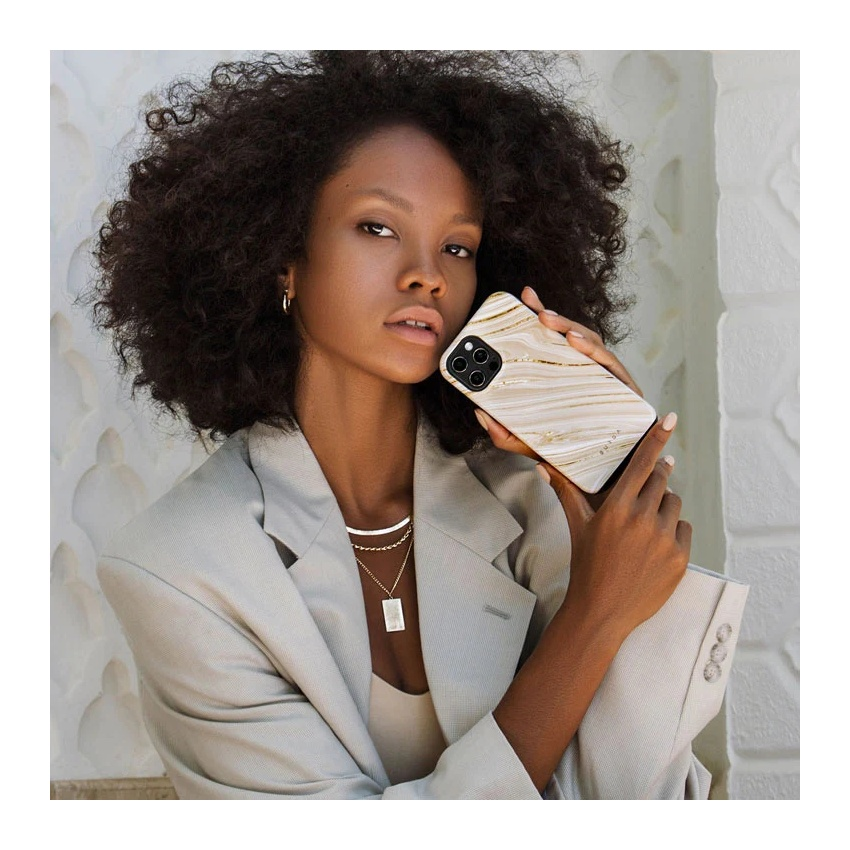 A woman with curly dark hair wearing a grey blazer holds a phone with a marble and gold case.