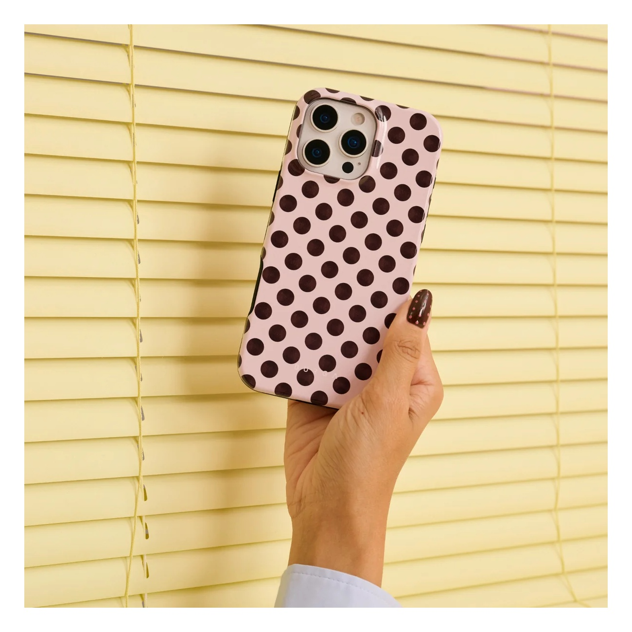 A hand holds a pink phone case with brown polka dots against yellow blinds.