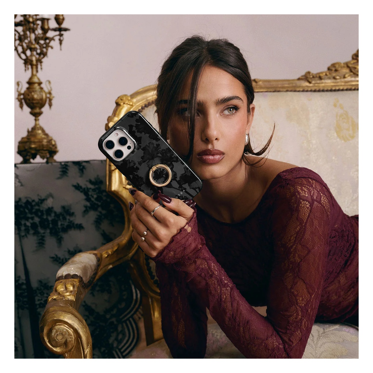 A woman in a maroon lace top holds a black phone case with a ring stand, seated on an ornate gold chair.