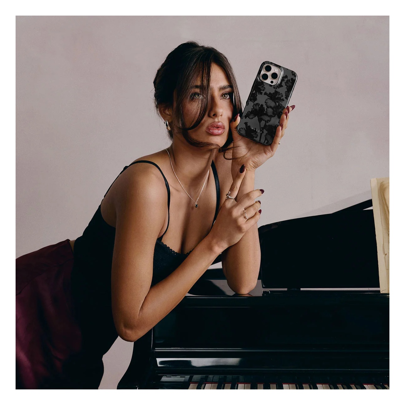 A woman leans against a black grand piano, holding a floral phone case next to her face in an indoor setting.