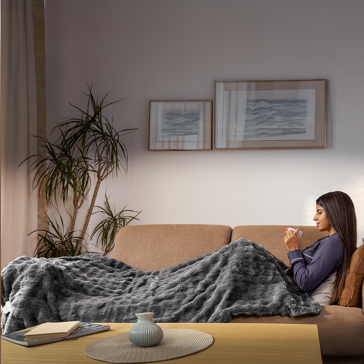 Una mujer se relaja en un sofá bajo una manta gris, sosteniendo una taza en una sala acogedora.