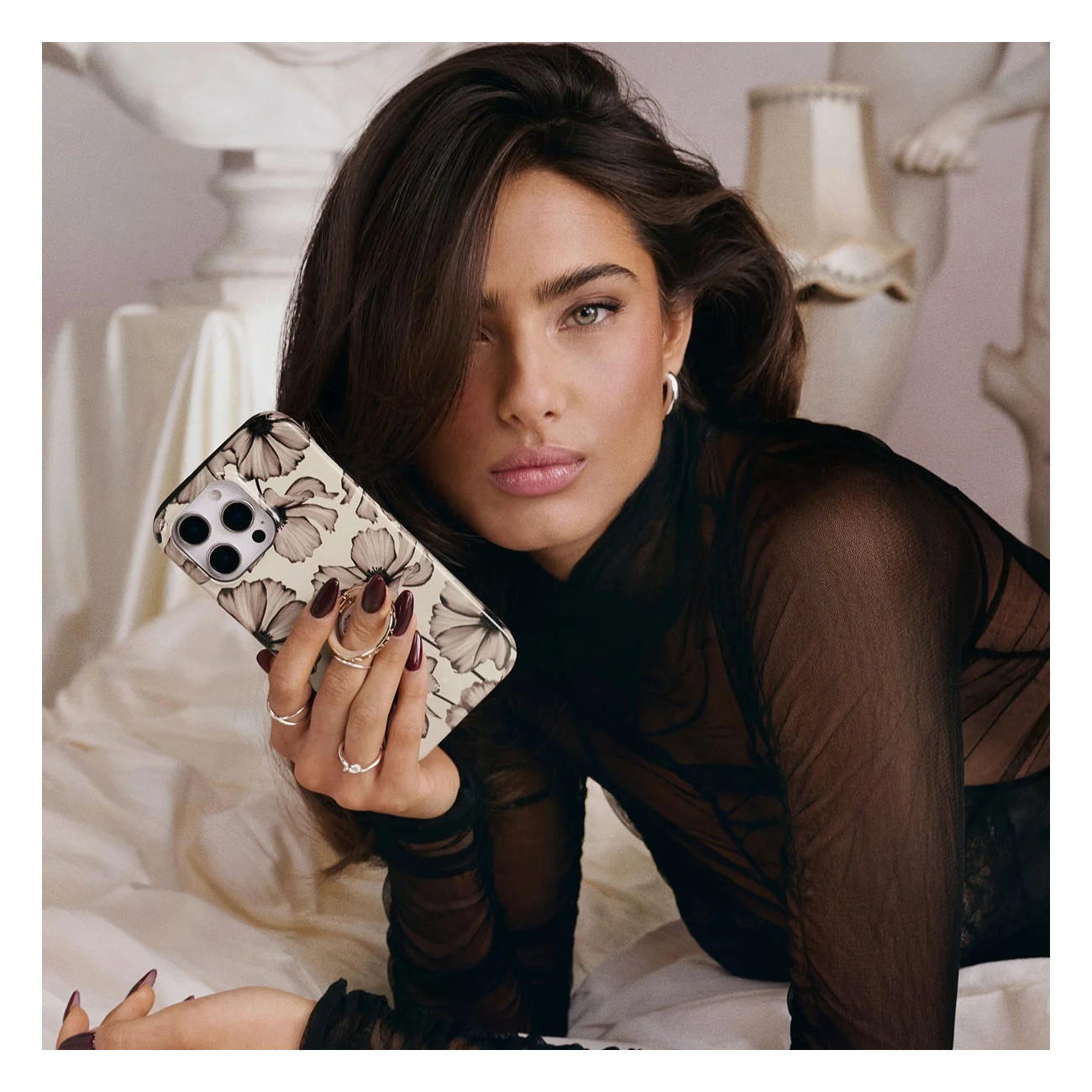 A woman with dark hair and rings holds a floral phone case, lying on a white surface with sculptures in the background.