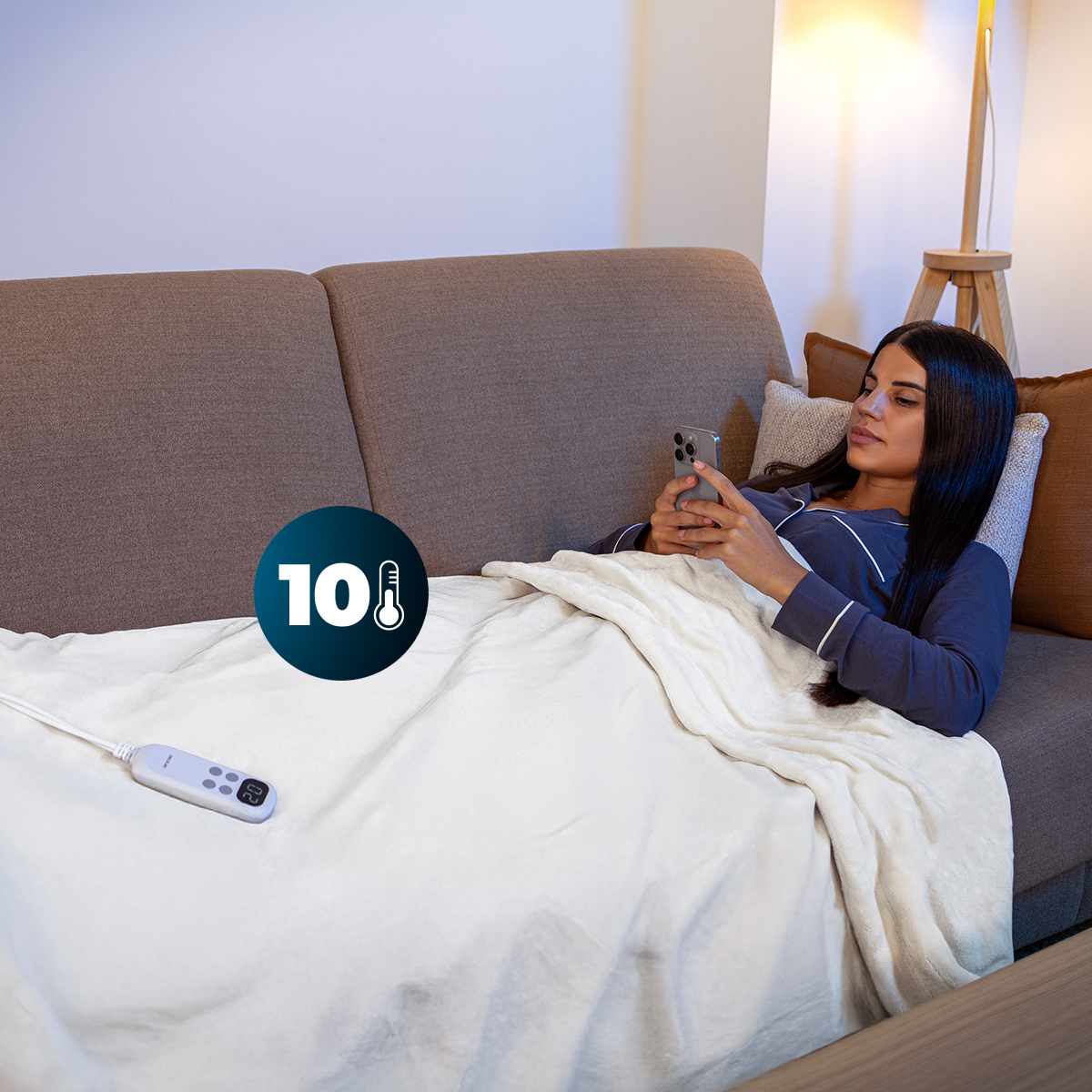 Una donna in pigiama blu è sdraiata su un divano, usa uno smartphone sotto una coperta bianca con un telecomando.