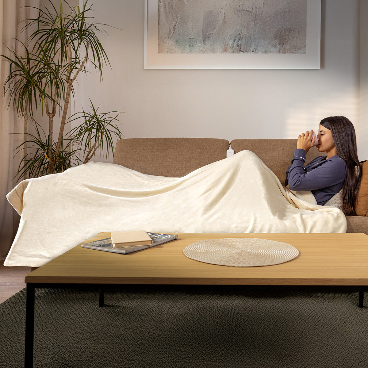Una donna si rilassa su un divano marrone sotto una coperta crema, bevendo da una tazza.