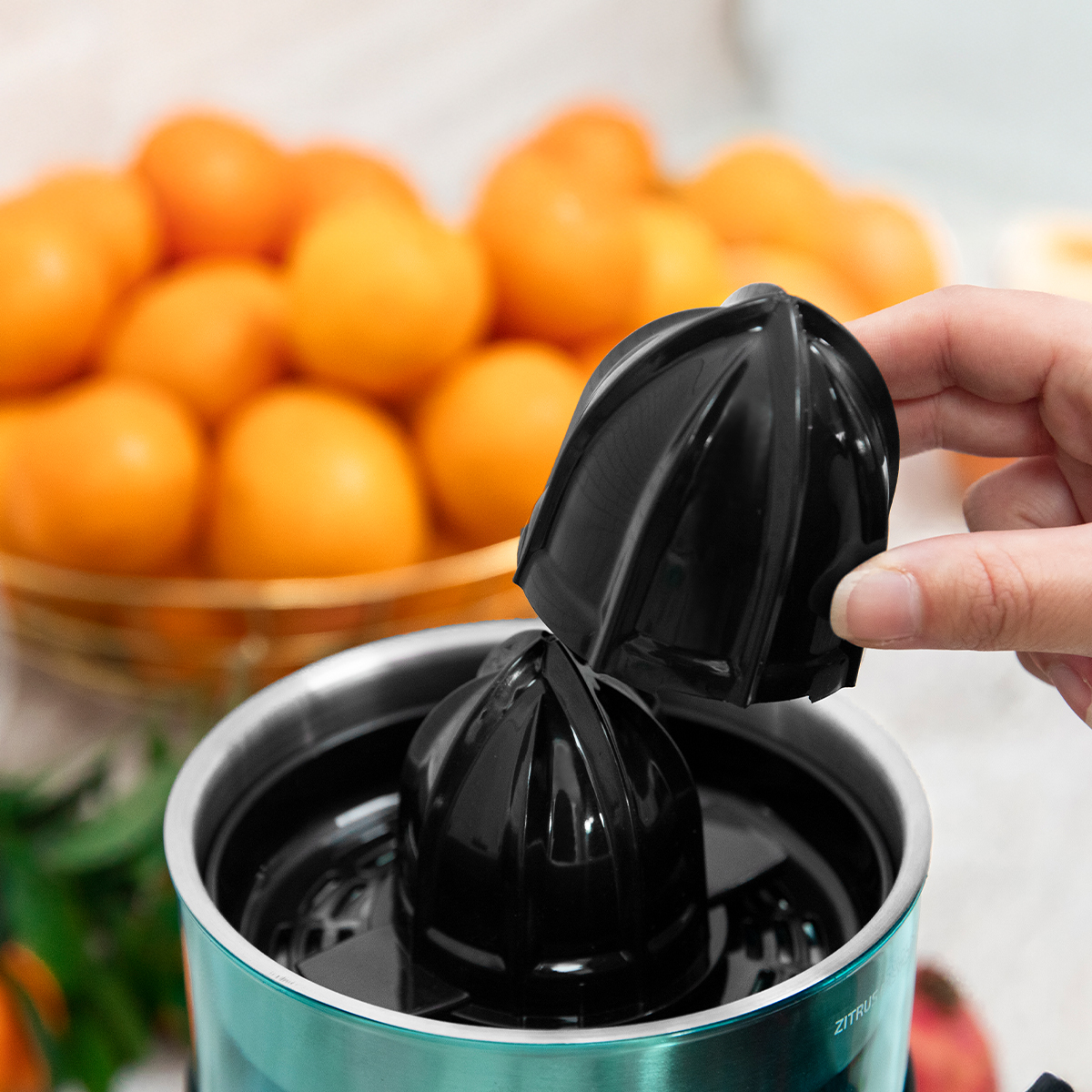 Una mano sostiene un accesorio exprimidor negro sobre un exprimidor eléctrico. De fondo, una cesta con naranjas.