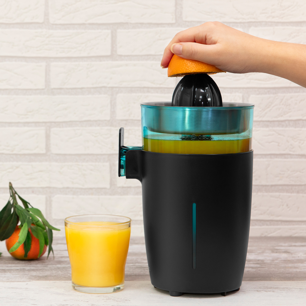 Una mano exprime media naranja en un exprimidor eléctrico negro junto a un vaso de jugo de naranja.