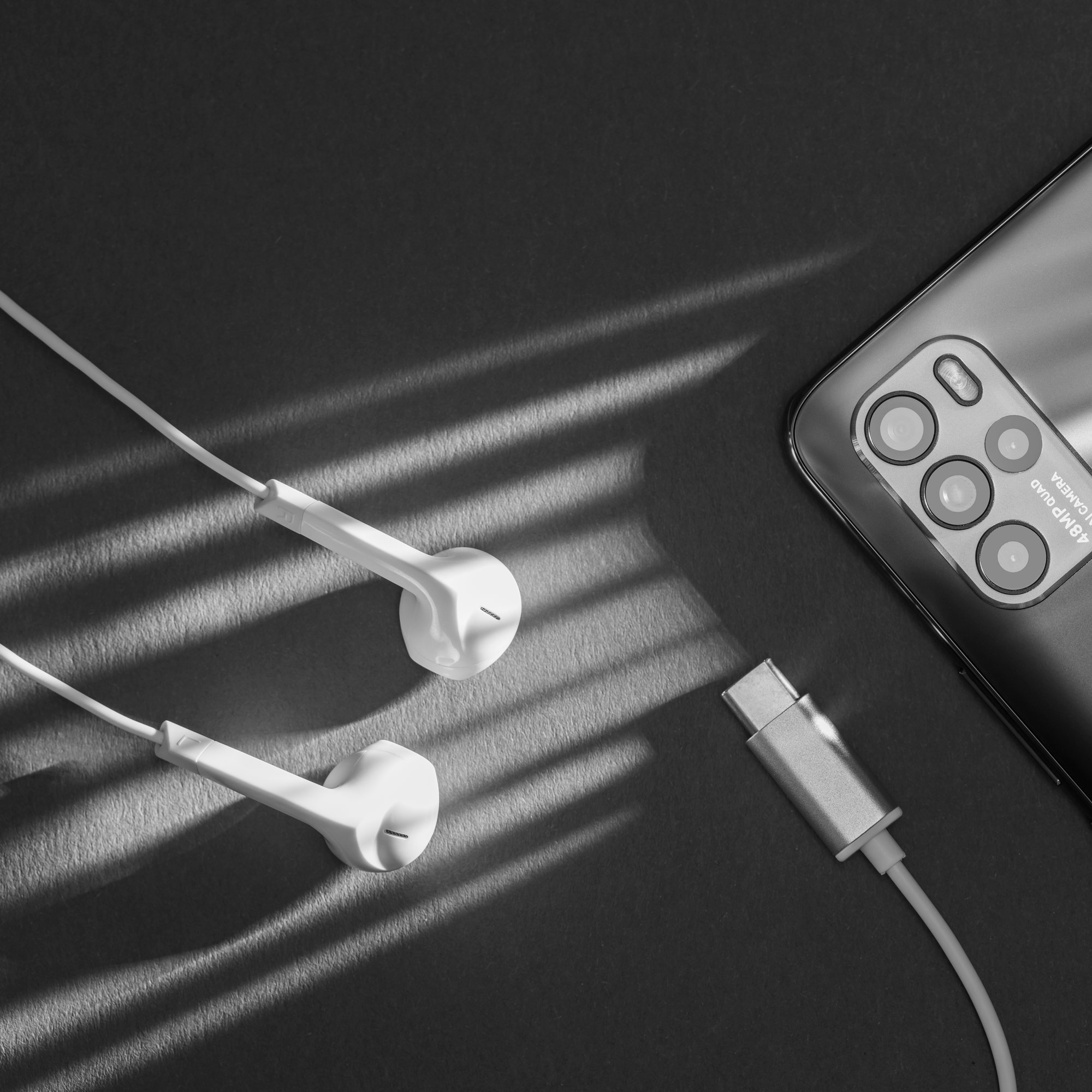 Białe słuchawki przewodowe, złącze USB-C i aparat w smartfonie na ciemnej powierzchni z pasami światła.