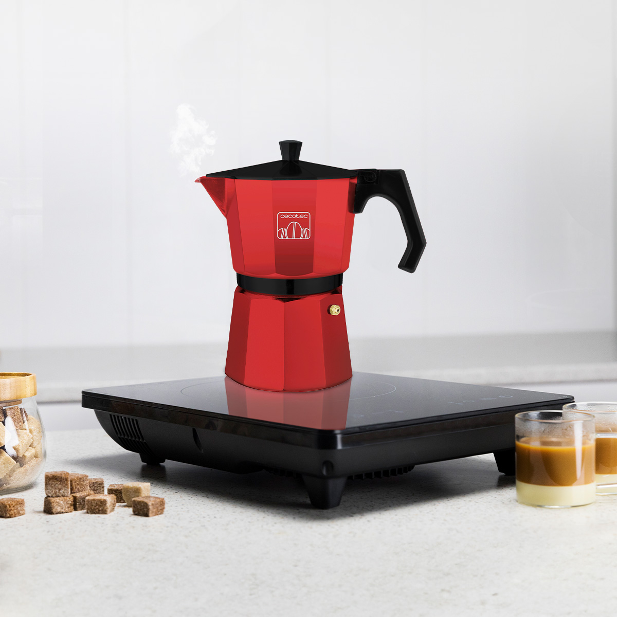 Una moka rossa su piano cottura a induzione nero, con vapore, zucchero in cubetti e due bicchieri di caffè.