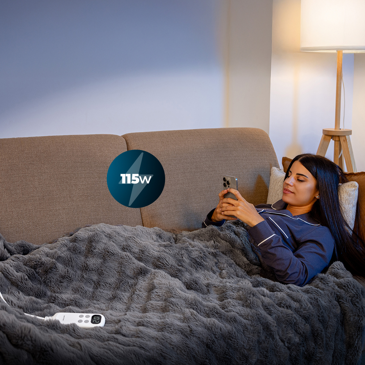 Una mujer en pijama gris se relaja bajo una manta suave en el sofá, mirando su teléfono inteligente.