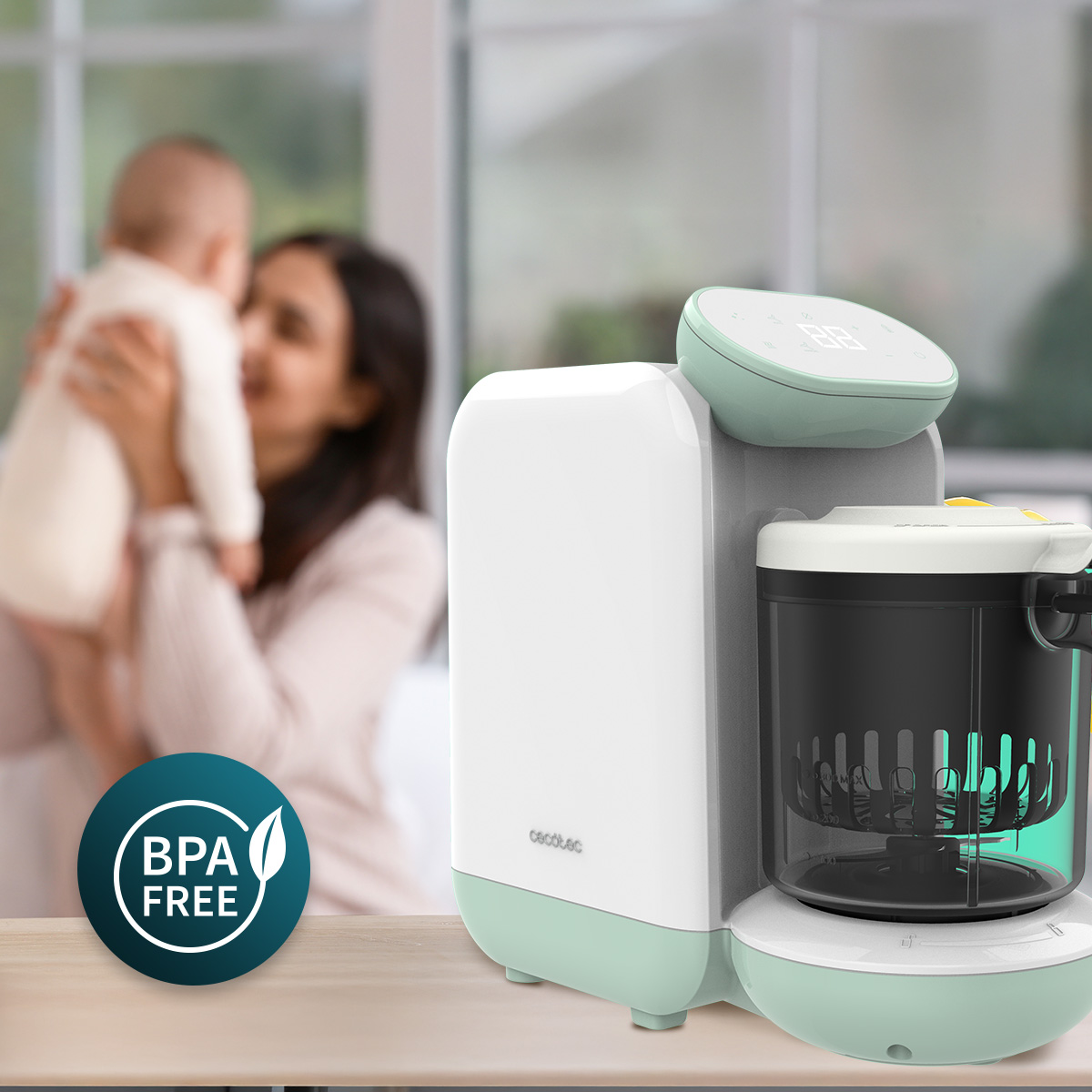 Un robot da cucina per neonati bianco e verde menta su un tavolo, con una madre che tiene il suo bambino sullo sfondo.