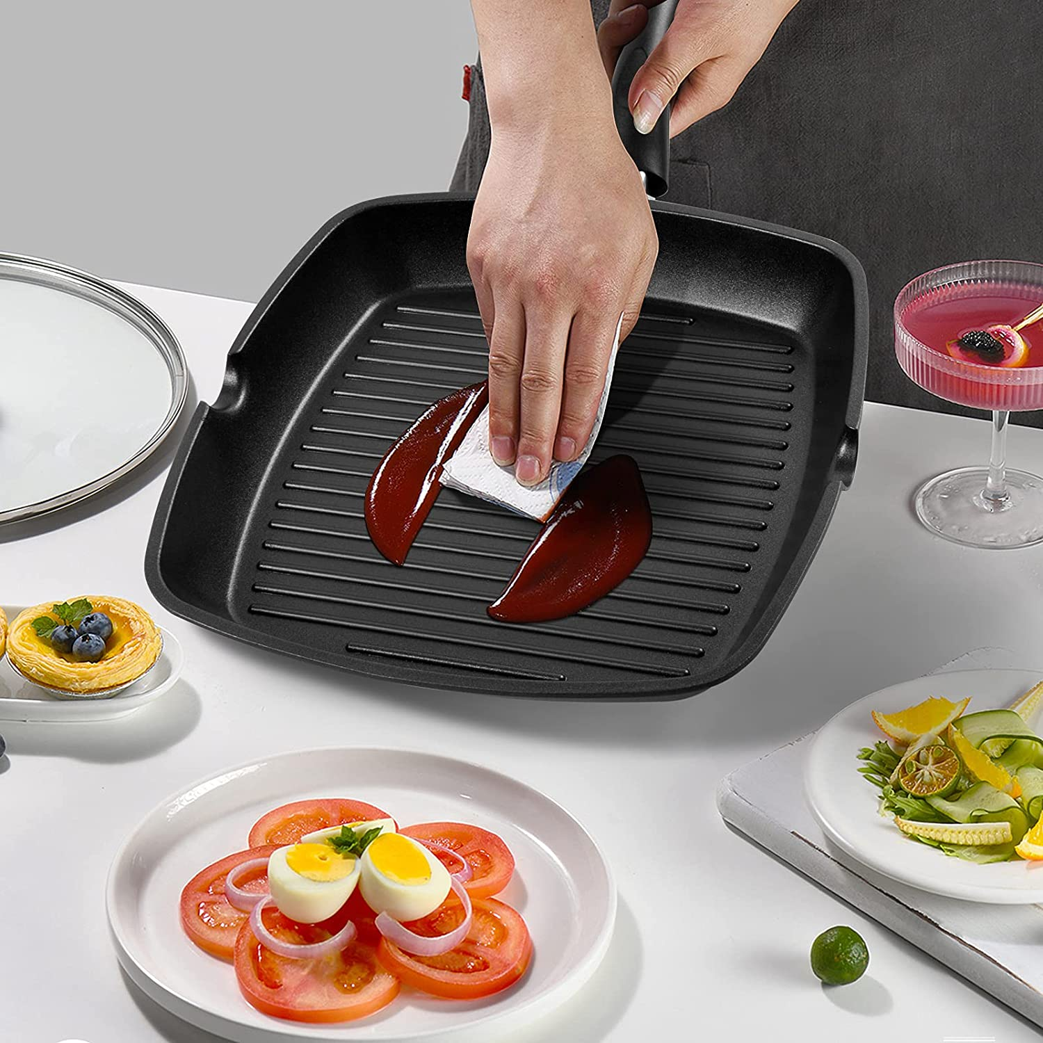Eine Person wischt eine schwarze Grillpfanne mit einem Papiertuch aus, umgeben von Salat, Eiern und einem Getränk.