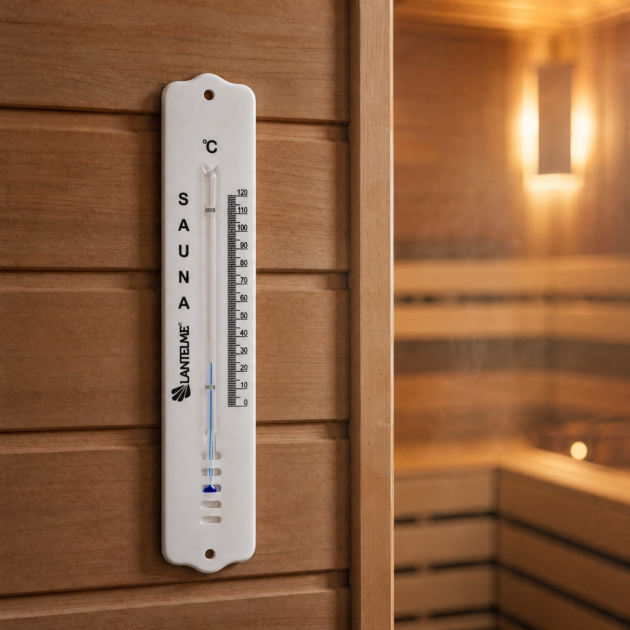 Ein weißes Saunathermometer hängt an einer HolzWand, im Hintergrund ist ein heller, warmer Saunaraum zu sehen.
