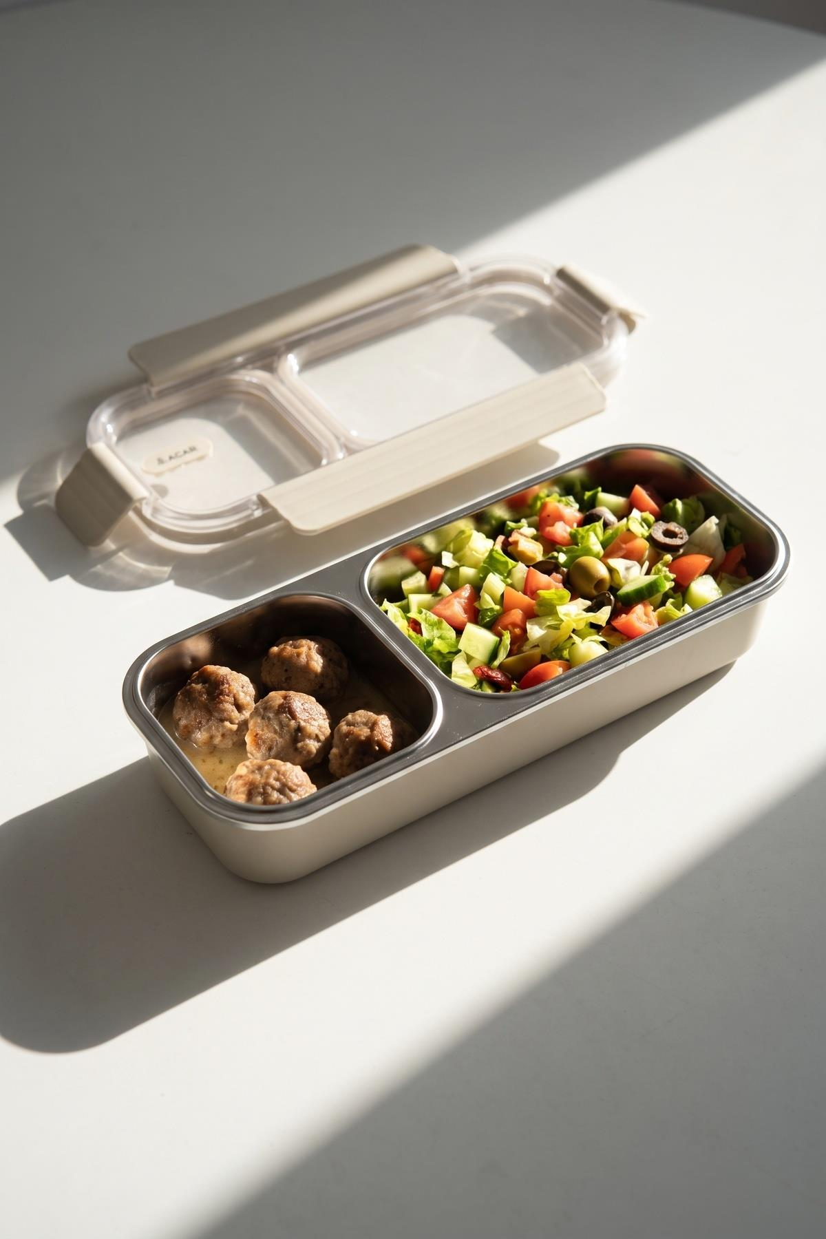 Bölmeli bir beslenme çantasında soslu köfte ve domates, salatalık ve zeytinli bir salata var.