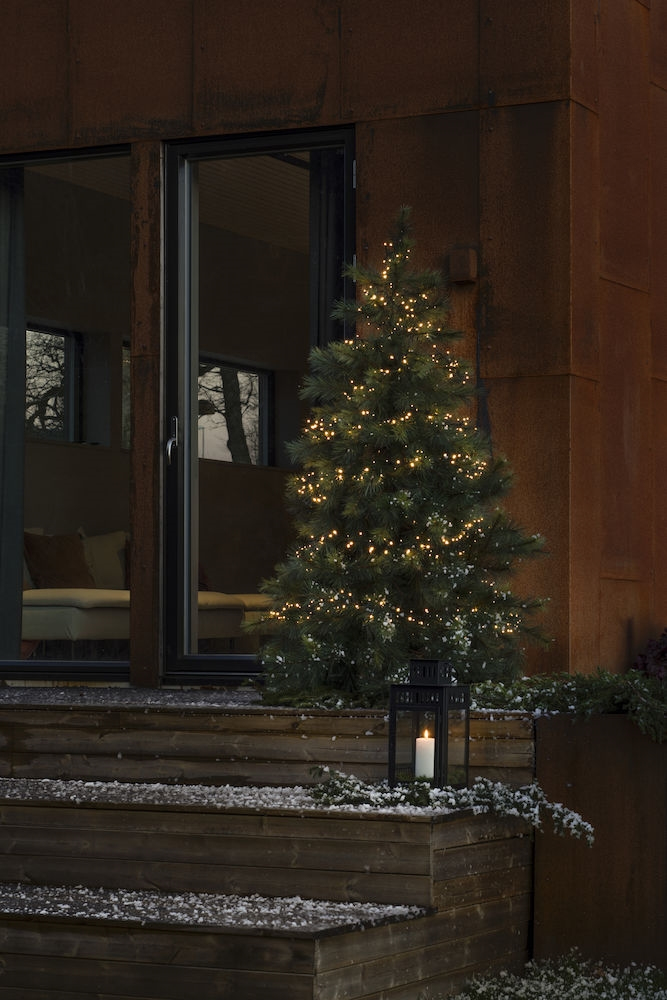 Ein beleuchteter Weihnachtsbaum auf Holzstufen vor einem rostigen Metallhaus mit Kerzenlaterne.