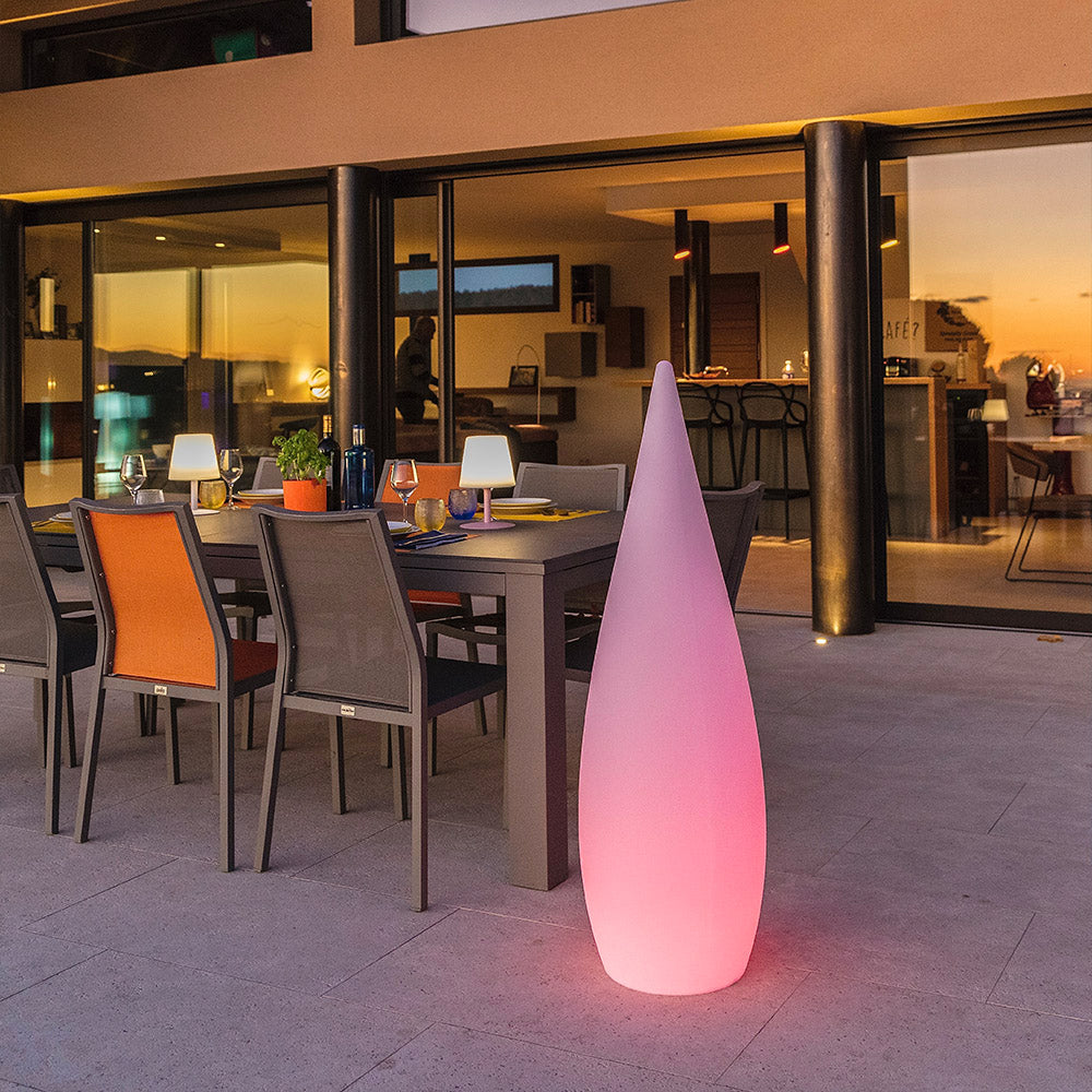 Eine rosafarbene, tropfenförmige LED-Lampe steht nachts auf einer Terrasse vor einem beleuchteten Essbereich.