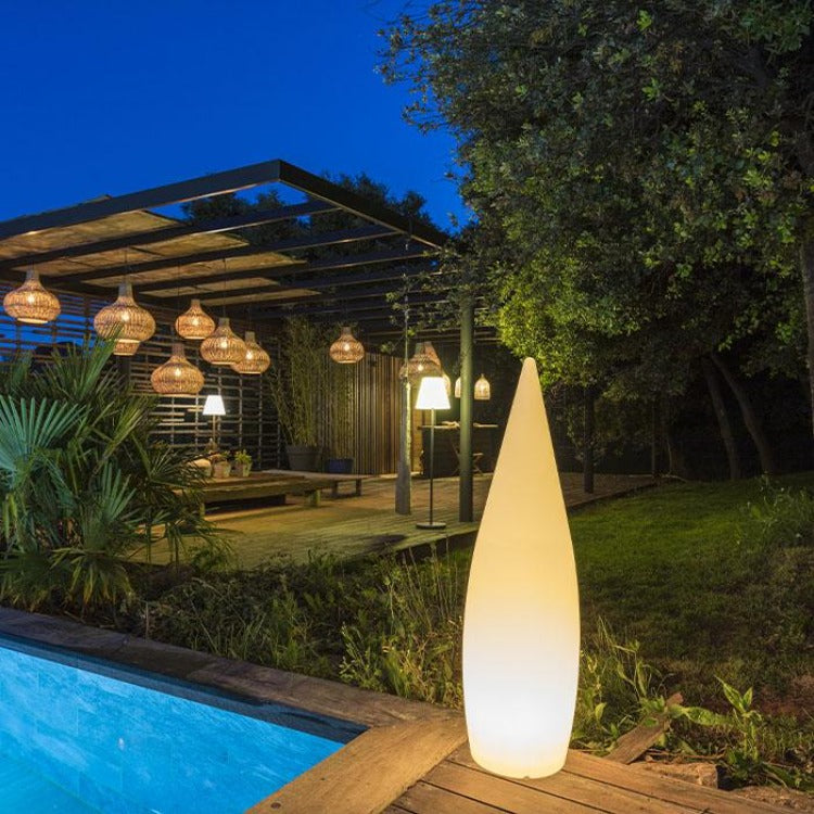 Beleuchtete kegelförmige Lampe am Poolrand bei Nacht, im Hintergrund eine Pergola mit Pendelleuchten und Bäumen.