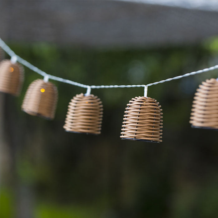 Eine Lichterkette mit braunen, geflochtenen Lampenschirmen hängt draußen im Garten.