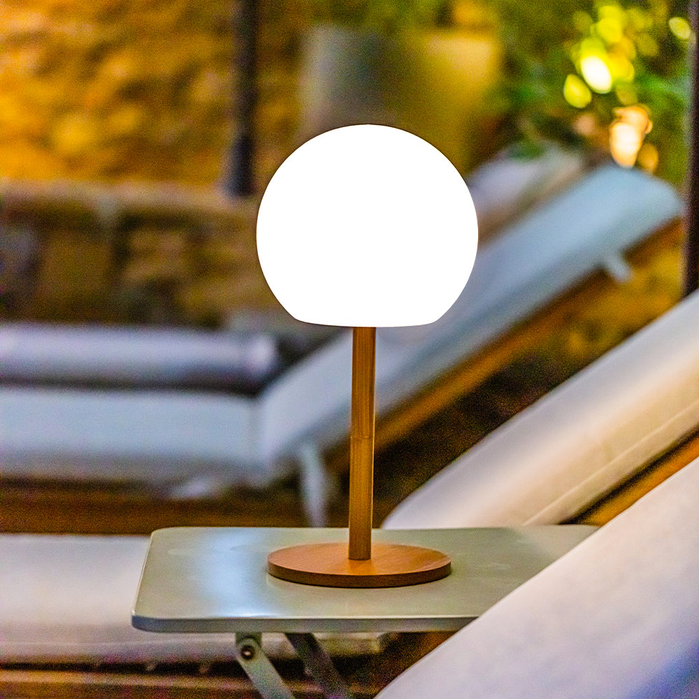 Eine runde, leuchtend weiße Lampe auf einem Holzständer steht im Freien auf einem kleinen Tisch.