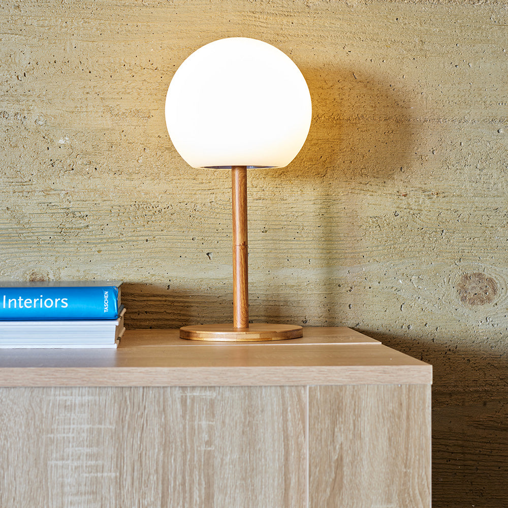 Eine runde weiße Tischlampe mit Holzfuß steht auf einem Holztisch vor einer strukturierten Wand.