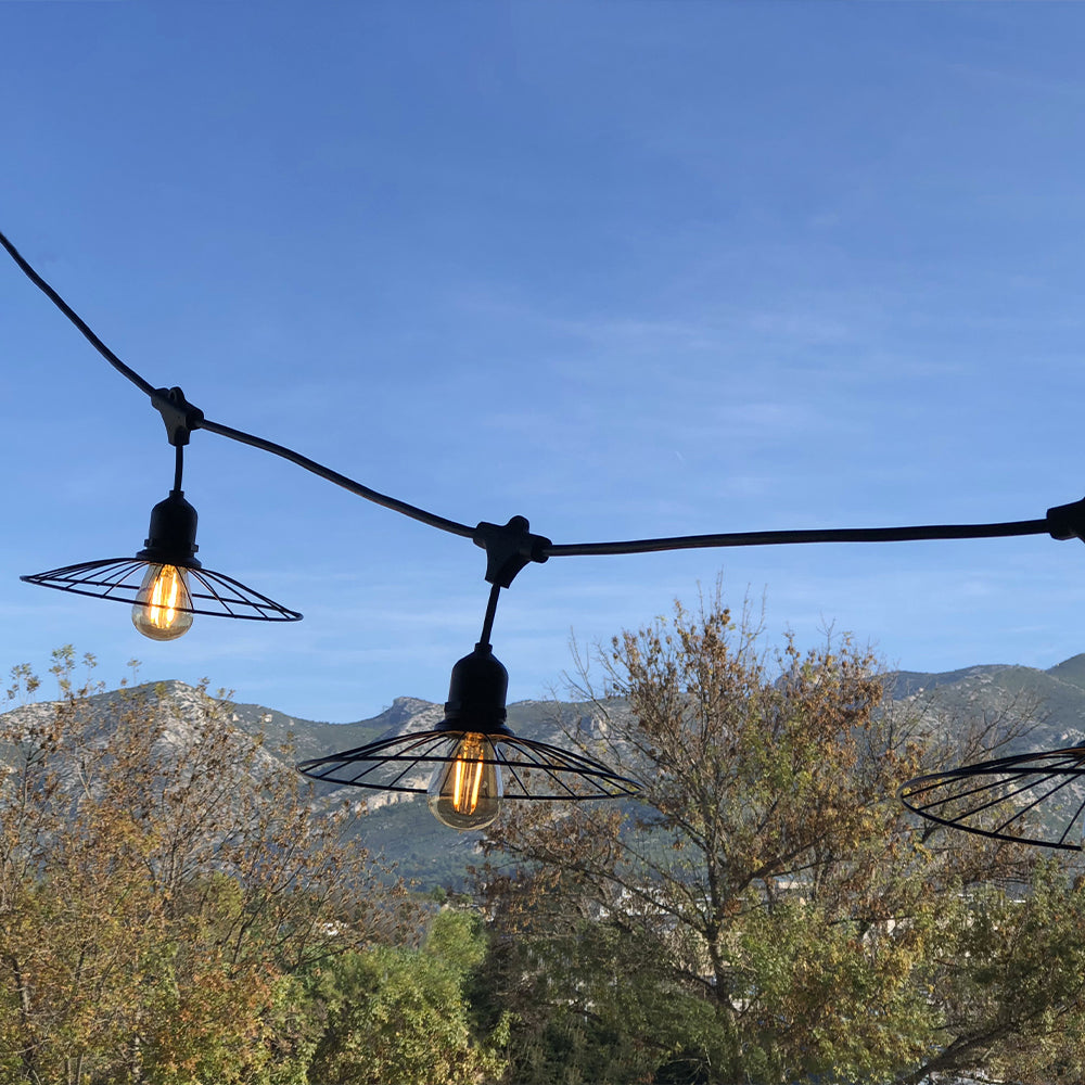 Lichterkette mit Drahtschirmen hängt vor blauem Himmel, Bäumen und Bergen.