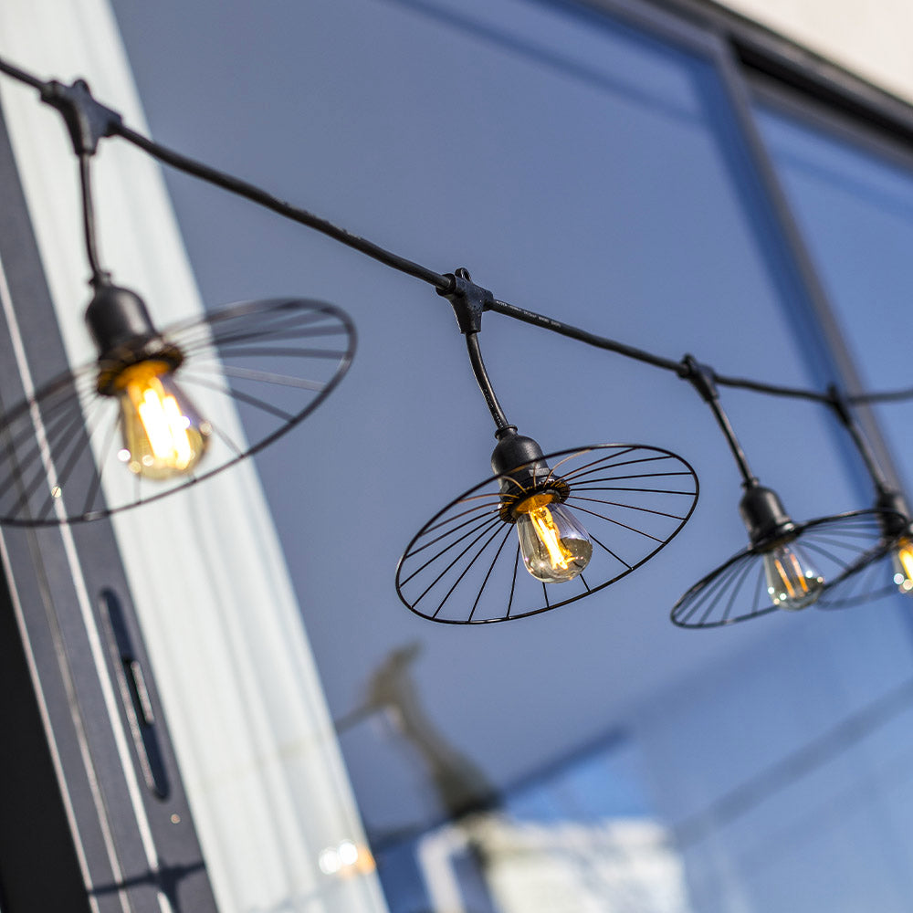Lichterkette mit Edison-Glühbirnen und schwarzen Drahtschchirmen vor blauem Glas.