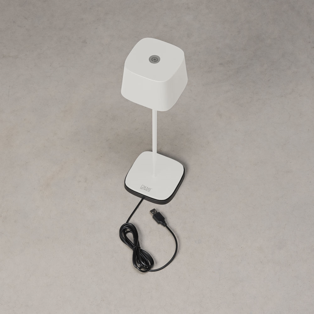 Eine weiße Tischlampe mit quadratischem Schirm und Fuß steht auf grauem Grund, ein schwarzes USB-Kabel liegt davor.