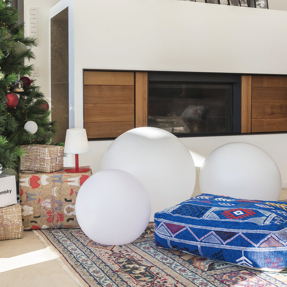 Wohnzimmer mit Weihnachtsbaum, Geschenken, beleuchteten Kugellampen, Kamin und blauem Musterkissen.
