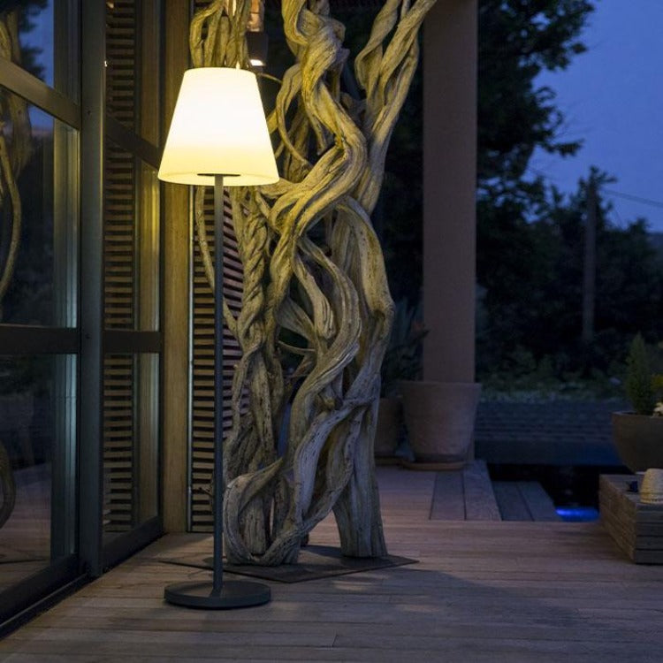 Lampada da terra illuminata su terrazza in legno al crepuscolo, con scultura di rami sullo sfondo.